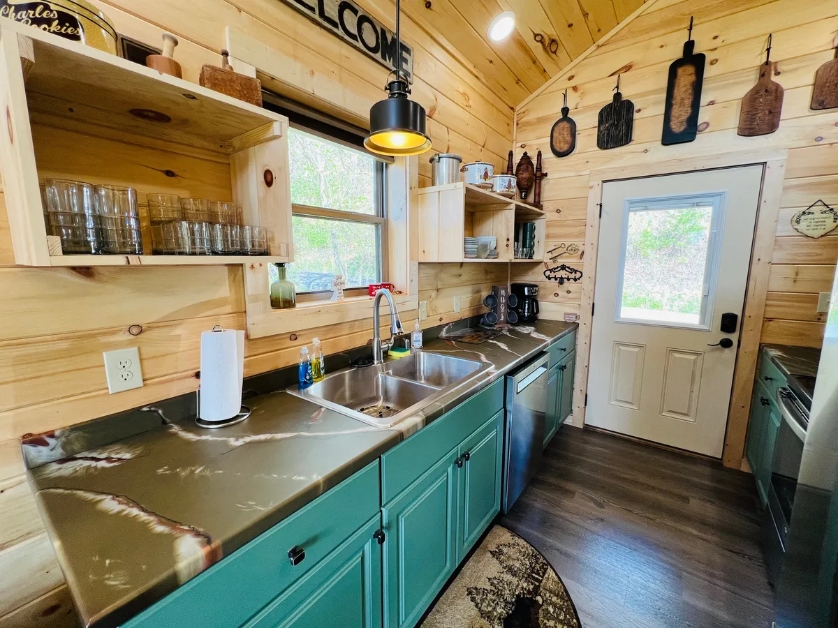 An inviting kitchen area with a kitchen sink, countertop, open shelves, and a welcoming sign, all bathed in natural light from a window. The space feels cozy and functional, ideal for gathering and enjoying morning coffee or meal preparation.