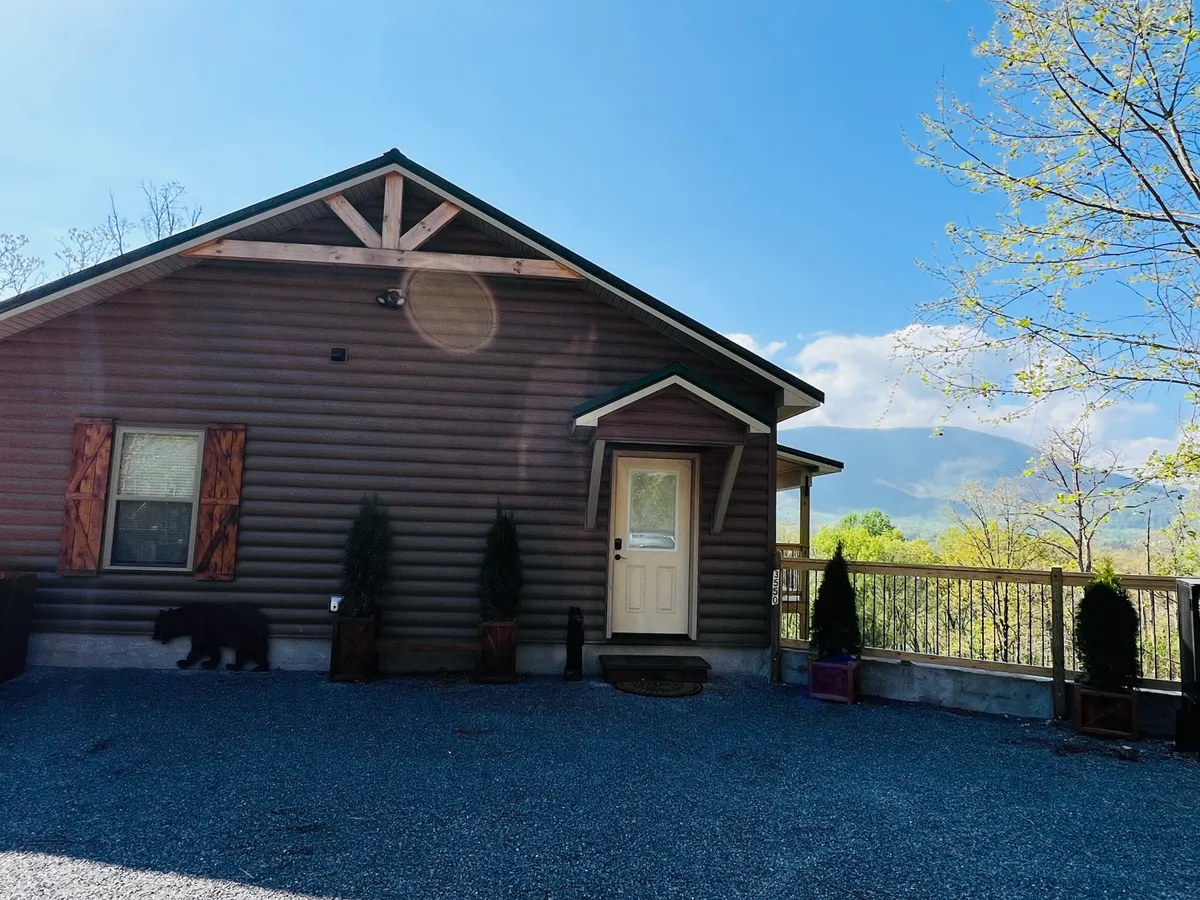 A view of the log cabin exterior surrounded by lush greenery, with wooden shutters, garden planters, and a welcoming doorway. The natural setting is serene and the cabin appears nestled in the wilderness, inviting guests to step outdoors and explore.