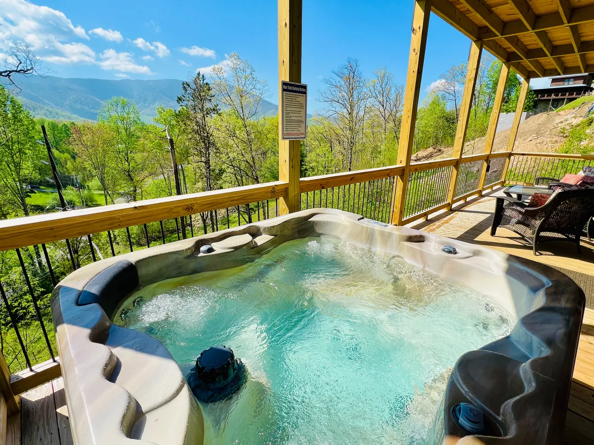 On the spacious wooden deck, a bubbling hot tub overlooks picturesque mountain views. A nearby seating area offers a spot to relax and take in the scenery, blending indoor comfort with outdoor serenity.