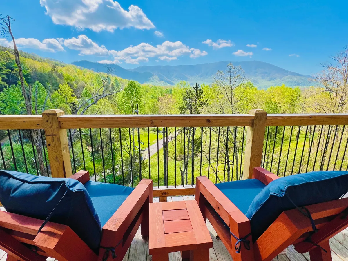 A tranquil exterior shot showcasing a wooden deck with Adirondack chairs and a stunning mountain view. Lush greenery surrounds the seating area, providing a peaceful setting for socializing, stargazing, or enjoying the sunrise together.