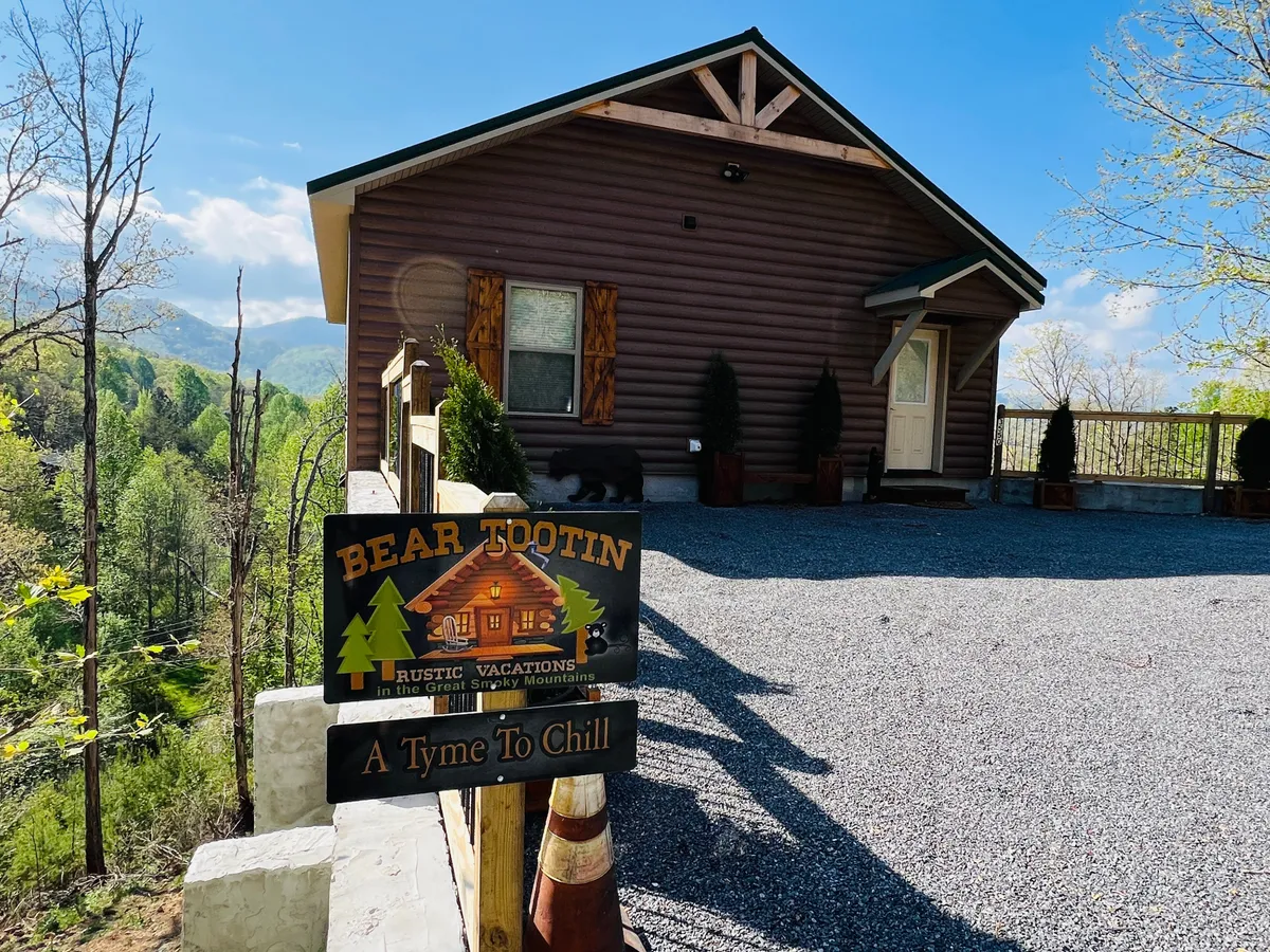 An inviting exterior view of the cabin's facade, featuring the 'Bear Tootin'' sign, warm wooden tones, and a mountain backdrop. The porch promises serene sunset views and a welcoming arrival.