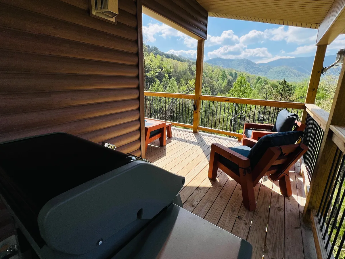 A spacious wooden deck furnished with lounge chairs and a grill, set against a backdrop of mountains. The space is designed for relaxation, outdoor dining, and socializing while enjoying the natural beauty that surrounds the cabin.