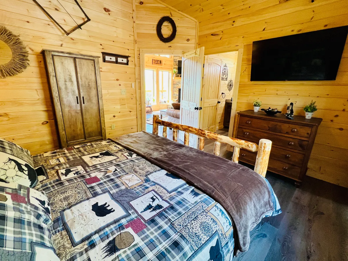 A second king bedroom with a wooden bed frame and plaid bedding. The room includes a wooden wardrobe, TV mounted on the wall, and decorative pieces on a dresser. The overall setting radiates comfort and relaxation, offering a homey yet upscale vibe suitable for unwinding after adventure-filled days.