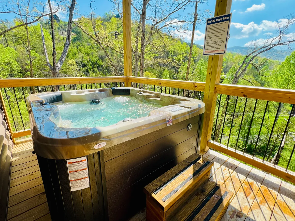 An outdoor hot tub set on a wooden deck surrounded by lush nature, with ample space for relaxing and socializing. The deck offers panoramic mountain views, ideal for soaking after a day of adventure or stargazing in the evening.