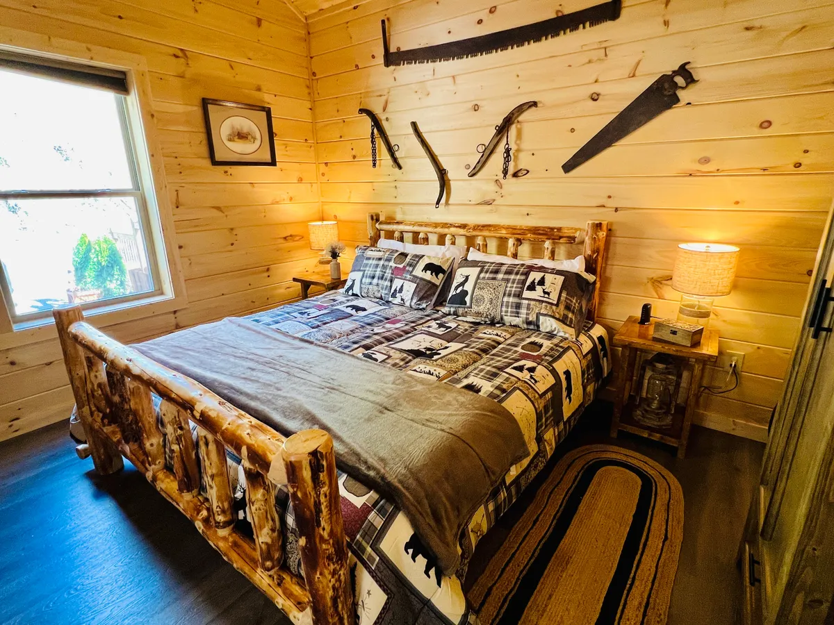 A cozy master bedroom featuring a rustic log king bed adorned with a colorful patchwork quilt. The room is accented by wooden walls, vintage tools as decor, a window letting in natural light, and a bedside table, creating a warm and inviting atmosphere that feels both luxurious and tranquil.
