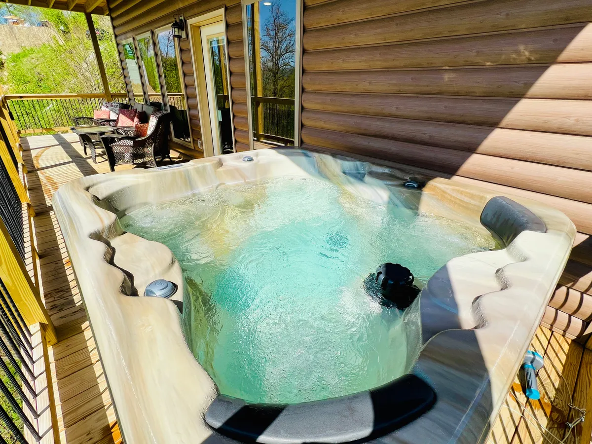 This image captures a wooden deck with a hot tub and outdoor seating area, set against the backdrop of a classic log cabin. The deck is spacious with comfortable seating, inviting guests to soak in the hot tub while taking in the beautiful natural views, or to gather for conversations and relaxation outdoors.