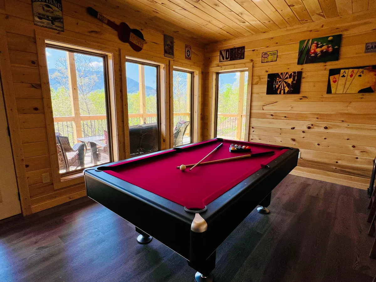 This image captures a spacious game room centered around a pool table, with large windows along the wall revealing scenic outdoor views. The wooden walls and decorative wall art give the room a warm, rustic cabin feel. It embodies an atmosphere of fun, relaxation, and togetherness, with room for socializing and friendly competition.