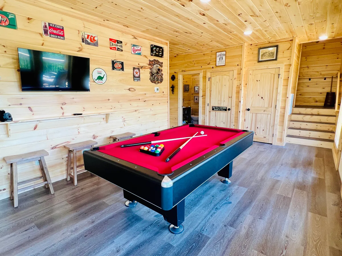 A spacious, fun-filled game room featuring a pool table, wall-mounted television, wooden bar stools, and playful signage. The space is designed for family game nights, movie watching, and social gatherings, with a casual and inviting atmosphere.