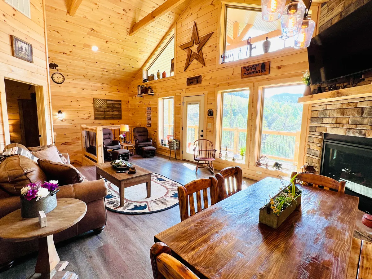 A spacious and sunlit open-concept living and dining area, featuring large picture windows with sweeping mountain views. The room boasts a cozy stone electric fireplace, a wooden dining table with seating for six, plush seating including a comfortable sofa, and rustic accents throughout. Natural light fills the space, creating a warm and welcoming atmosphere.
