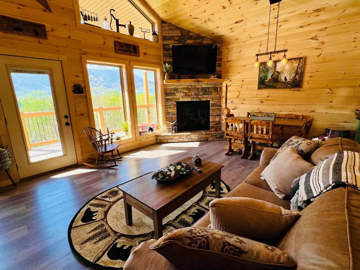 A warmly decorated living and dining area with expansive windows showcasing scenic mountain views. The stone fireplace anchors the space, while a wooden dining table and inviting sofa offer comfortable spots for relaxation or shared meals. The decorative rug and soft lighting add to the cozy, communal vibe.