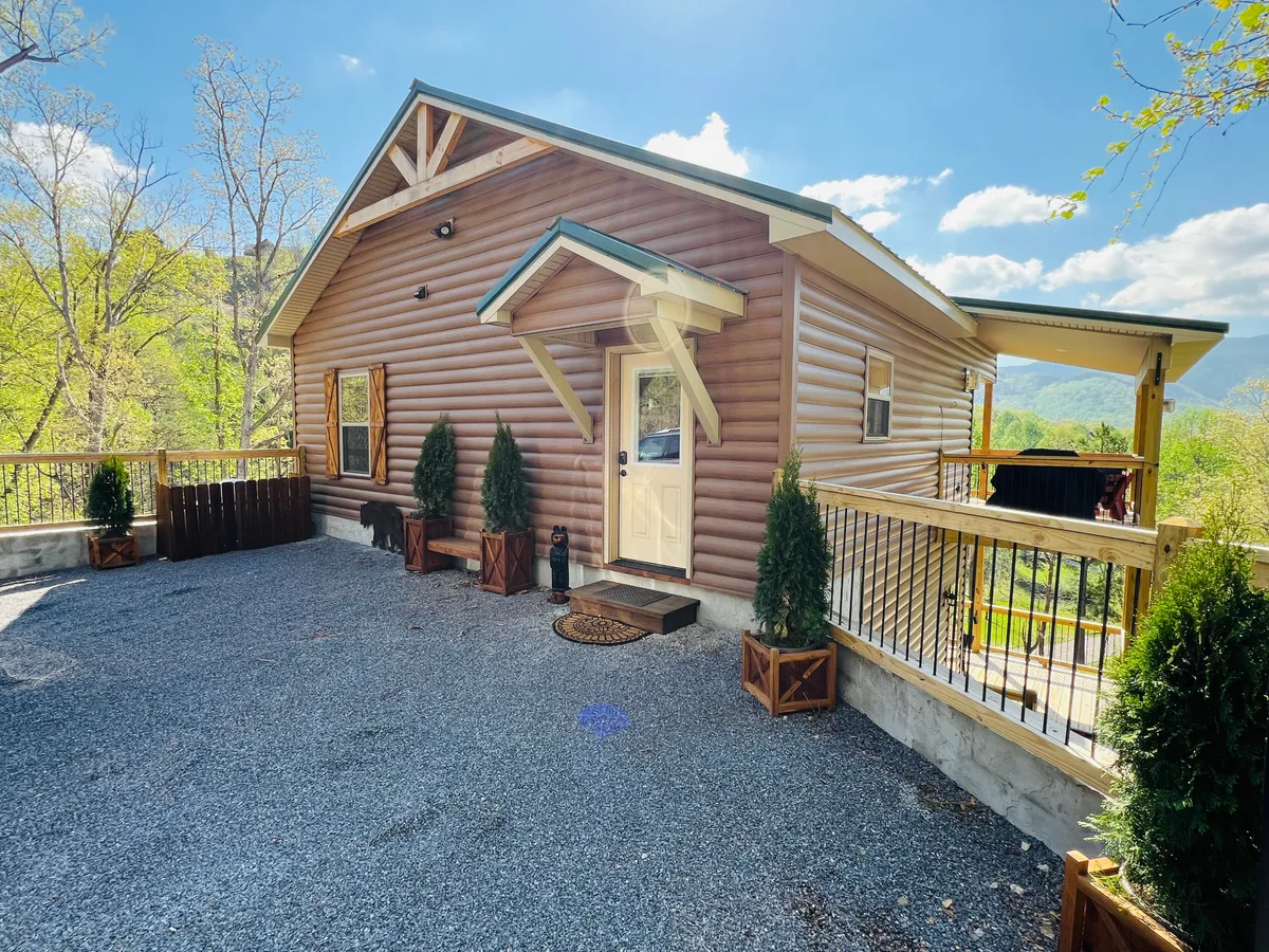 A welcoming exterior view of the log cabin, showcasing its distinctive architecture, a spacious porch with inviting seating, and well-maintained landscaping set against a backdrop of natural greenery. The image captures the essence of arriving at a serene mountain retreat.